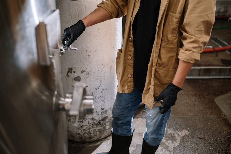 A brewmaster wearing gloves operates a beer tap in an industrial brewery setting.